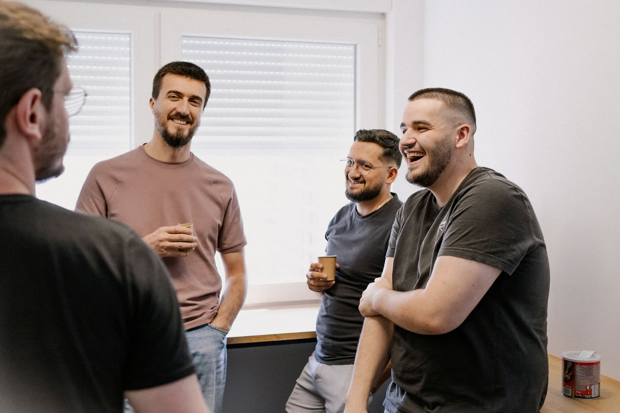 Four employees of a small business laughing together over coffee during a break in a warm, sunlit office