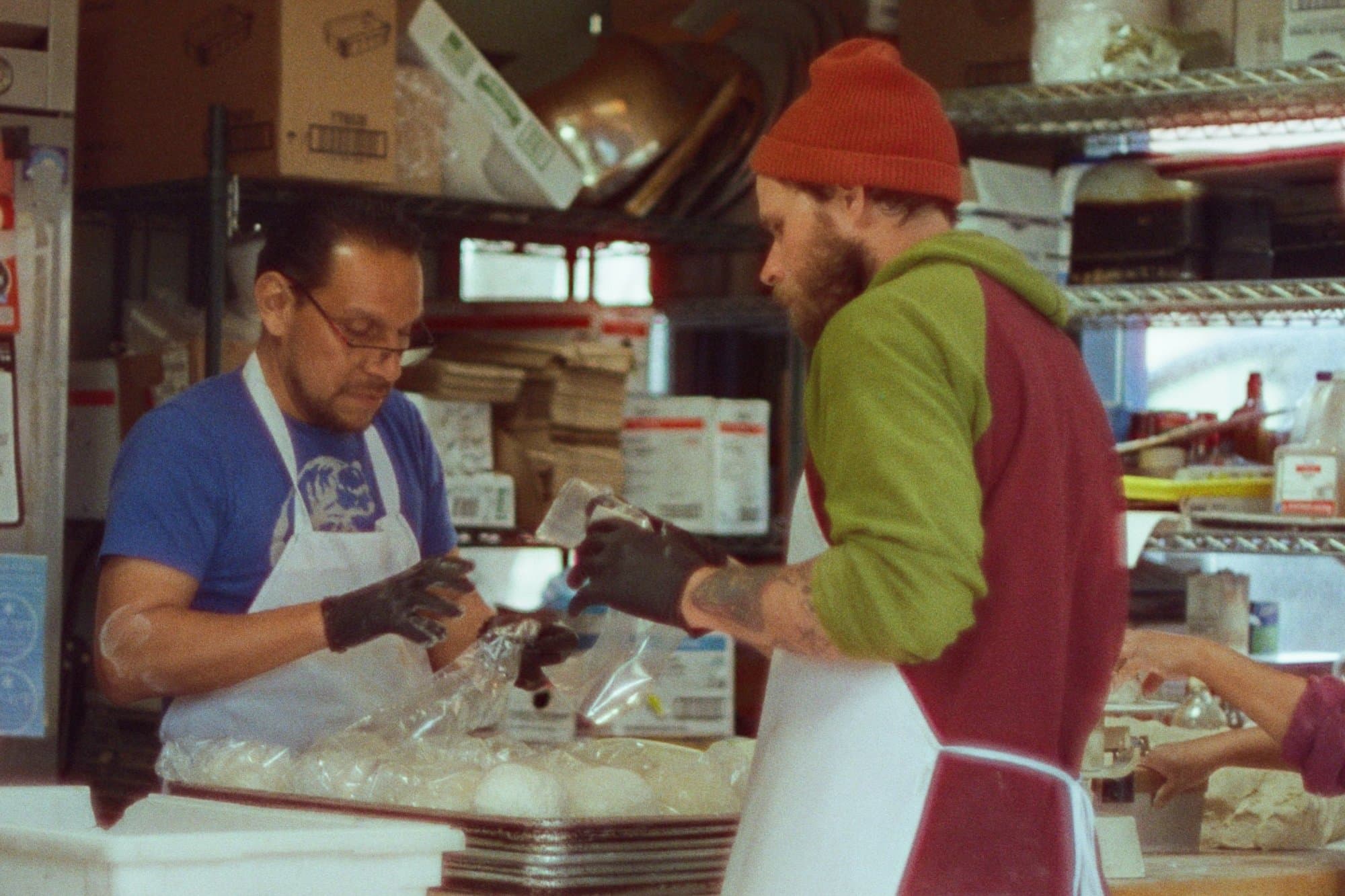 Two aproned bakery workers prepping dough side by side at the table of a small bakery kitchen